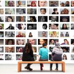 Drei Personen sitzen auf einer Bank und betrachten eine große Wand mit zahlreichen Porträtfotos.